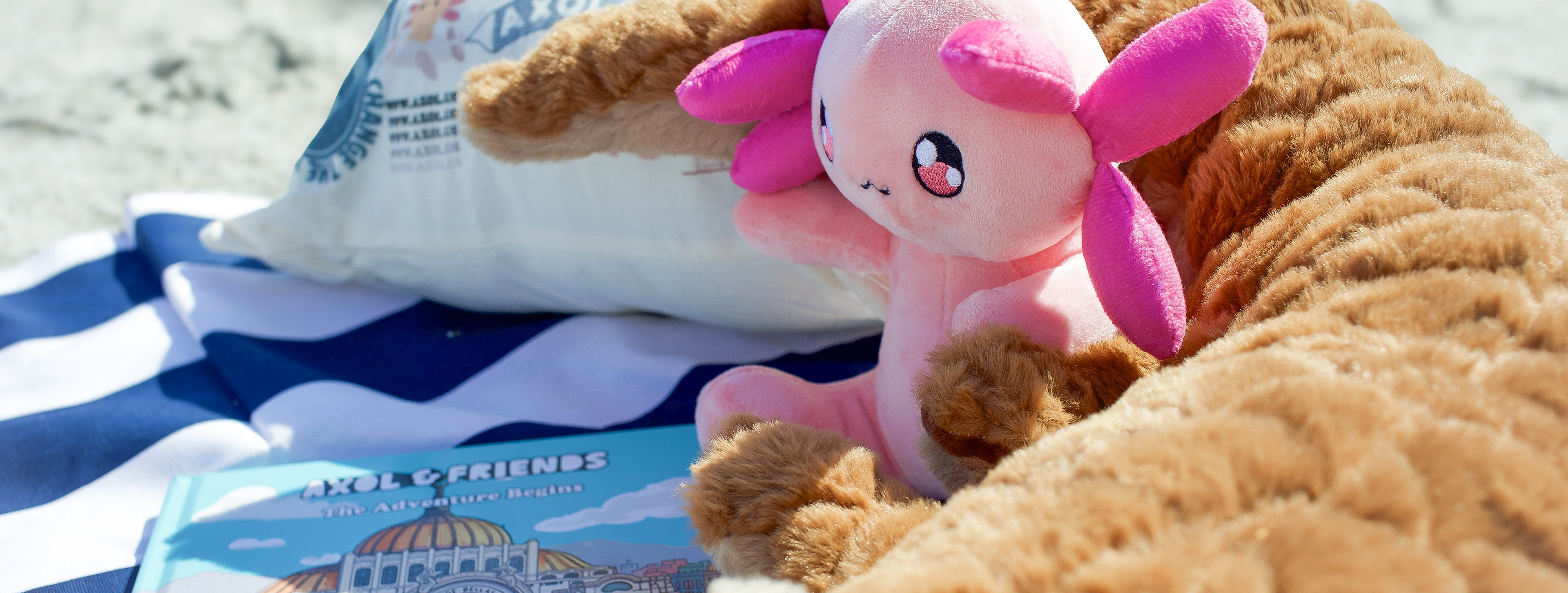A pink plush toy axolotl sits on a striped beach towel next to a white pillow and a blue book on a sandy beach.