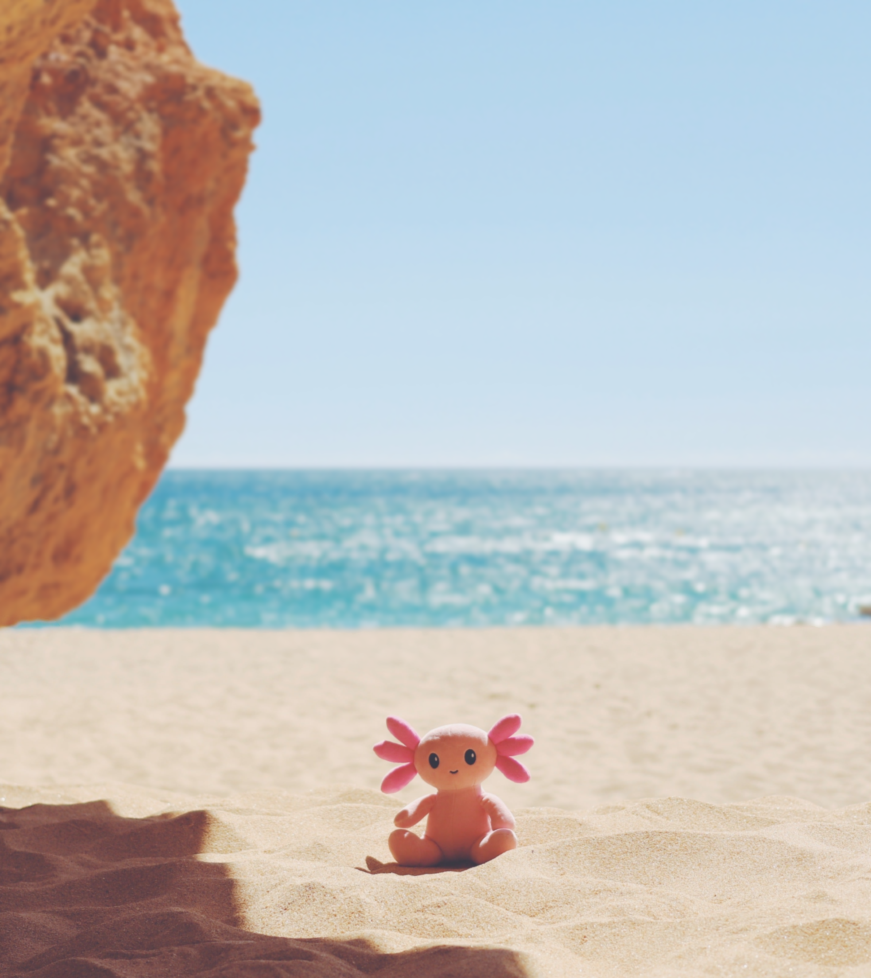 A small pink and orange axolotl-shaped plush toy sits in the sand on a beach, with a large rock on the left, blue ocean water in the background, and a pale blue sky above.